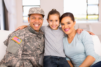 Soldier in military fatigues sitting on a couch with his wife and daughter smiling at the camera